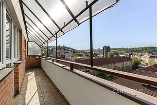 Prostorný balkon s dlažbou nabízí panoramatický výhled na město, doplněný částečně zastřešenou konstrukcí a cihlovým obložením.