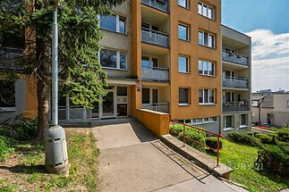 Vchod do bytového domu se širokou přístupovou cestou, moderní fasádou a balkóny. Nachází se v klidné lokalitě s upravenou zelení.