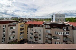 Pohled na městskou zástavbu s moderními bytovými domy, červené střechy, velká okna, výhled na park a vzdálené panorama, vhodné pro klidné bydlení.