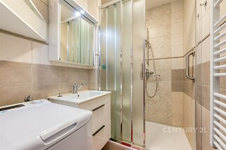 Malá koupelna s moderními obklady, sprchovým koutem a pračkou. Světlé dlaždice a zrcadlo vytváří pocit prostornosti.