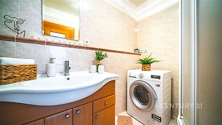 Kompaktní koupelna s moderními obklady, pračkou a dřevěnými úložnými skříňkami, nabízí praktické využití prostoru a světlý design.