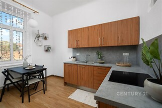 Moderní kuchyň s dřevěnými skříňkami, parketovou podlahou a jídelním koutem. Světlý interiér s velkým oknem a minimalistickým designem.