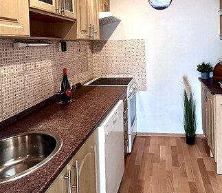 Úzká kuchyň s dřevěnými skříňkami, moderními spotřebiči a parketovou podlahou, nabízí útulný prostor pro vaření.