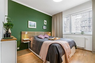Světlá ložnice s parketovou podlahou, moderním nábytkem a velkými okny s žaluziemi, nabízí útulný prostor k odpočinku.