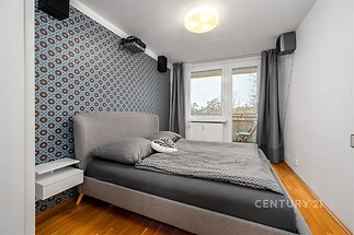 Stylová ložnice s dřevěnou podlahou, velkými okny a moderní tapetou. Světlý a útulný prostor ideální pro relaxaci.