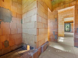 Nedokončený interiér domu s cihlovými zdmi, betonovou podlahou a velkými okny, které zajišťují světlost a potenciál pro moderní přestavbu.