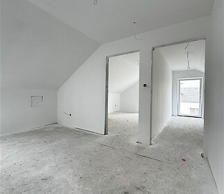 Podkrovní místnost s bílými stěnami a šikmým stropem, nedokončená podlaha. Velká okna zajišťují světlost, prostor pro vestavěnou skříň.