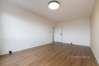 Prázdný pokoj s moderní parketovou podlahou, bílými stěnami a výrazným stropním svítidlem, ideální pro přizpůsobení osobním potřebám.