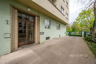 Vstupní prostor bytového domu s velkými okny, betonovou podlahou a rekonstruovanou fasádou, obklopený zelení a bezpečným oplocením.