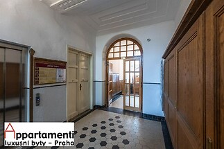 Chodba historického domu s dlažbou, dřevěným obložením a prosklenými dveřmi, které zajišťují vstup do dalších částí budovy.