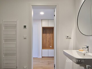 Moderní koupelna s bílými stěnami, kulatým zrcadlem, umyvadlem a designovým topným tělesem, parketová podlaha v předsíni s vestavěným nábytkem.