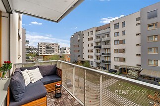 Balkon s dřevěnou podlahou a výhledem na moderní bytové domy. Pohodlné posezení a prostor pro relaxaci dosvědčují úroveň komfortu a stylu.