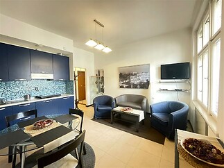 Kuchyňský kout s tmavě modrými skříňkami, světlá dlažba a moderní osvětlení, vedle obývací zóny s pohovkami a velkými okny.