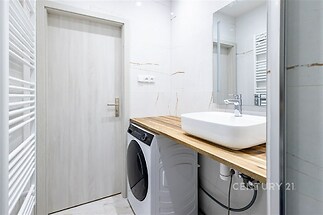 Koupelna s moderní bílou výbavou, dřevěnou pracovní deskou a vestavěnou pračkou, světlé dlažby dotvářejí čistý a elegantní vzhled.