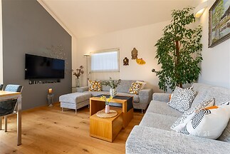 Světlý obývací pokoj s parketovou podlahou, moderní sedačkou a velkými okny. Dekorativní prvky a výborný stav vytvářejí útulný prostor.