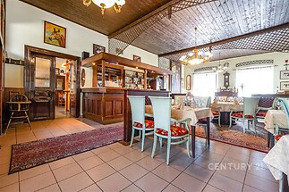 Restaurace v rustikálním stylu s dřevěným stropem, dlažbou a velkými okny zajišťujícími světlé prostředí, prostor je zařízený historickým nábytkem.