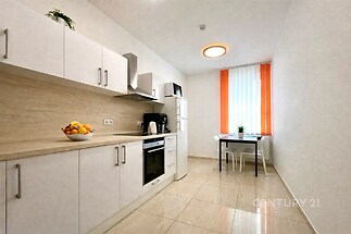 Moderní kuchyň s dlažbou, bílými skříňkami a vestavěnými spotřebiči, která má velká okna s oranžovými závěsy, poskytující dostatek světla.