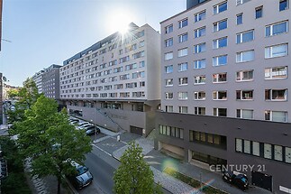 Moderní bytový dům s velkými okny, světlá fasáda a prostorná parkovací plocha, nachází se v klidné části města.