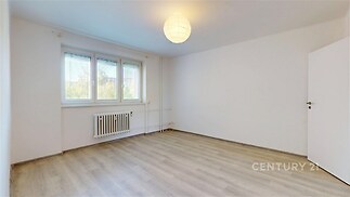 Prázdný pokoj s moderní laminátovou podlahou, bílými stěnami a velkými okny, zajišťujícími dostatek přirozeného světla.