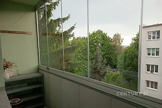 Zasklená lodžie s výhledem na zeleň, vysoké stromy a okolní budovy, útulný prostor, ideální pro odpočinek, klidné bydlení, moderní úprava