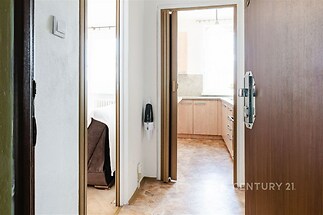 Vstupní chodba s dlažbou, ústící do světlé kuchyně s moderními skříňkami a dřevěnými povrchy, vedle ložnice s parketovou podlahou.