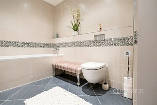 Moderní koupelna s dlažbou, světlé obklady a designový prvek na stěně, závěsná toaleta, stylová vana a dekorativní rostliny pro elegantní vzhled.