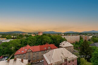 Výhled na město z vysokého patra zahrnuje střechy budov, zelené parky a panorama hor v pozadí, ideální pro milovníky přírody.