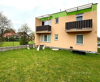 Zahrada s posezením u moderního domu, fasáda v pastelových barvách, malá balkony, udržovaný trávník, klidná rezidenční oblast.