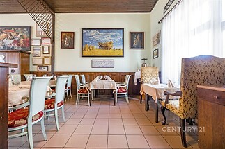 Restaurace s dlažbou, velkými okny a dřevěným obložením zdí, nabízející útulný interiér s malbami, světlé stoly a čalouněné židle.