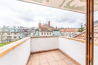Balkon s dlažbou a výhledem na historické budovy, částečně krytý, nabízí prostor k relaxaci v klidné lokalitě.