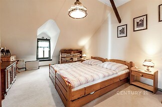 Podkrovní ložnice, dřevěná manželská postel, světlý koberec, dřevěné trámy, dekorativní osvětlení, útulný styl, decentní výzdoba na stěnách.