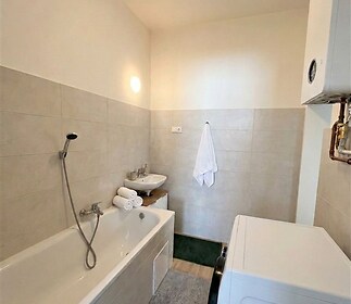 Koupelna s moderní dlažbou a světlými obklady, vybavená vanou, umyvadlem a pračkou.