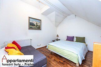 Podkrovní ložnice s dřevěnou parketovou podlahou, velkou postelí a světlými stěnami, nabízí útulný a moderní prostor pro relaxaci.