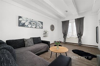 Světlý obývací pokoj s dřevěnou podlahou, velkými okny a moderním zařízením vytváří útulnou atmosféru pro relaxaci.