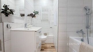 Moderní koupelna s bílými dlaždicemi, vanou a funkcionalistickým umyvadlem. Světlý interiér s minimalistickým designem a dřevěným prvkem.