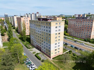 Venkovní pohled na panelový bytový dům v Praze s rekonstruovanou fasádou, parkovacími místy a blízkostí tramvajové zastávky.