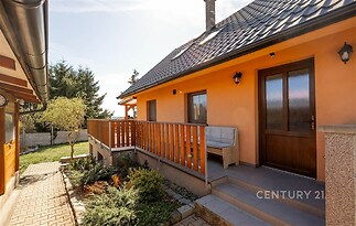 Fasáda rodinného domu s terasou, dřevěným zábradlím a moderními vstupními dveřmi. Udržovaná zahrada a dlážděná cesta doplňují venkovní prostor.