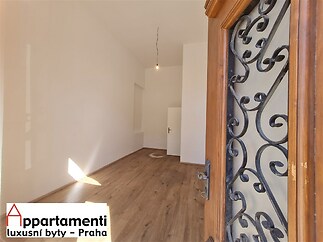 Úzká vstupní hala s parketovou podlahou a bílými stěnami, moderní vchodové dveře s ozdobným kováním, světlý interiér.