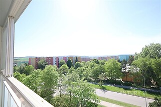 Výhled z balkonu na okolní zástavbu a zeleň, slunečný den s modrou oblohou, sídliště a stromy vytvářejí klidnou atmosféru.