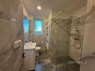 Moderní koupelna s velkoformátovou dlažbou, prosklený sprchový kout, velké okno, světlé povrchy z mramoru, moderní vybavení.