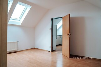 Podkrovní pokoj s laminátovou podlahou, střešní okna a bílými stěnami, moderní design, přístup do malé kuchyňky, světlý prostor.