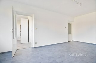 Světlý obývací pokoj s moderní laminátovou podlahou, čistě bílými stěnami a vstupem na chodbu, vkusně zrekonstruovaný prostor.