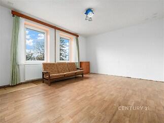 Prostorný obývací pokoj s dřevěnou podlahou, velkými okny a bílými stěnami nabízí světlý a vzdušný interiér vhodný k rekonstrukci.