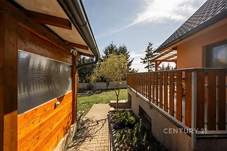 Terasa s dřevěným zábradlím u rodinného domu, přístupová cesta s dlažbou, dobře udržovaná zahrada, moderní venkovský styl.