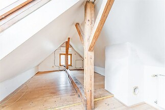 Podkrovní prostor s dřevěnou podlahou a trámy, světlý díky střešním oknům, ideální pro kreativní rekonstrukci nebo jako skladovací místnost.