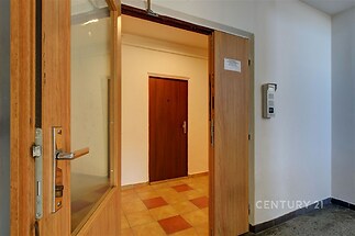 Vstupní hala s keramickou dlažbou, dřevěnými dveřmi a interkomem, ve světlém činžovním domě.