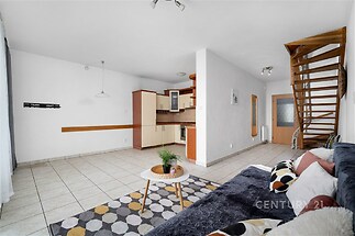 Obývací pokoj s moderní dlažbou, dřevěným schodištěm a světlou kuchyní. Velká okna umožňují přirozené světlo a vytváří prostorný dojem.