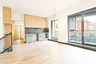 Světlý obývací pokoj s moderní kuchyní, parketová podlaha, velká okna a dřevěné detaily vytvářejí svěží atmosféru v novostavbě.