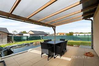 Zastřešená terasa s dlažbou, moderní zahradní nábytek u jezírka, výhled na klidnou obytnou čtvrť, ideální pro relaxaci a venkovní posezení.