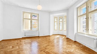 Světlý obývací pokoj s parketovou podlahou, velká okna nabízejí dostatek přirozeného světla, moderní styl s elegantními detaily.